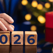 Hand adjusting wooden blocks to display “2026” with festive lights and holiday gift boxes in the background.